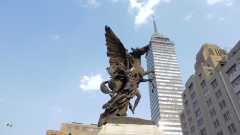 Eine-Weite-Zeitlupenaufnahme-Mit-Heranfahren-Eines-Pegasus-Und-Eines-Engelsdenkmals-Im-Vordergrund-Und-Des-Latinoamericana-Tower-Aus-Mexiko-Stadt-Im-Hintergrund-An-Einem-Klaren,-Blauen-Tag