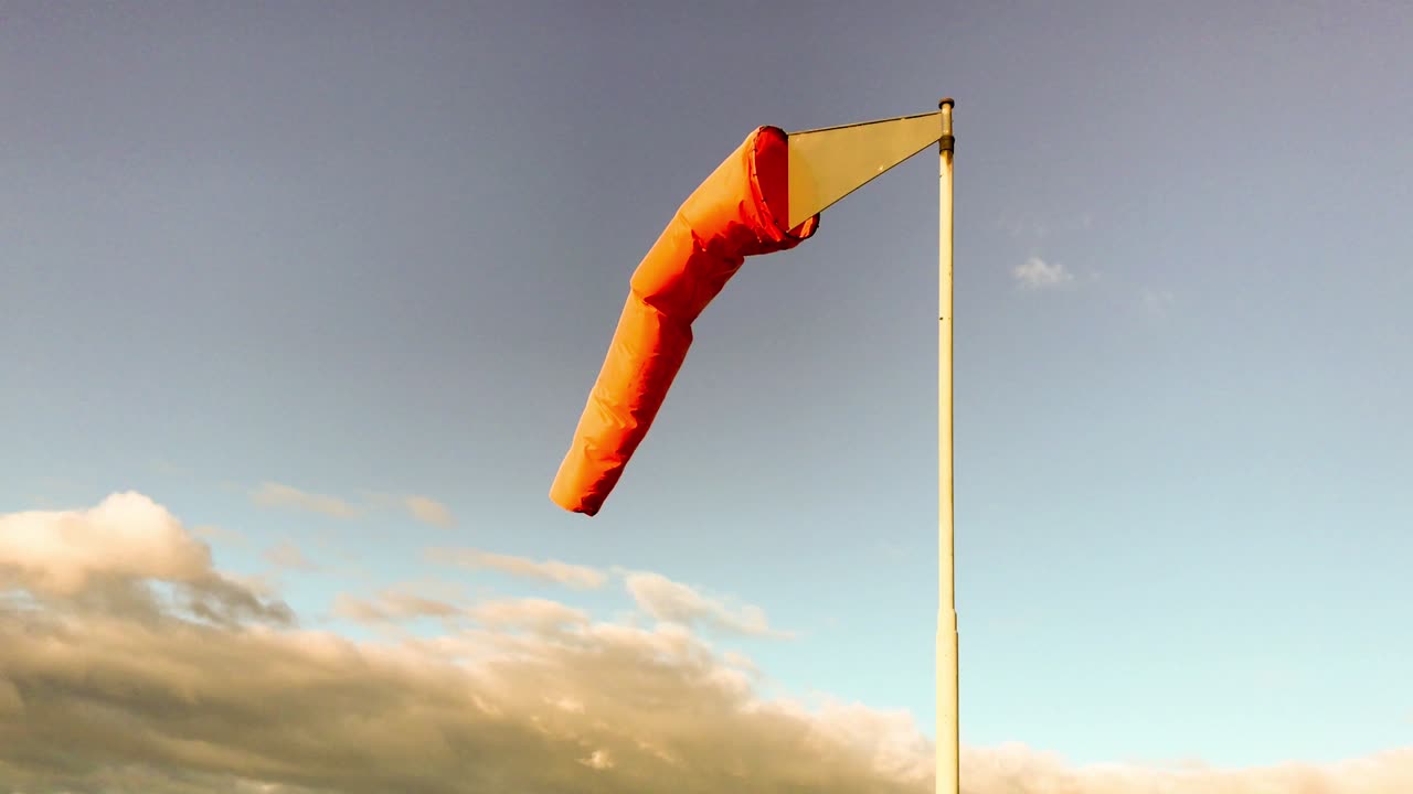 un windsock de color naranja sopla suavemente en el viento bajo un cielo azul con la aparición de nubes oscuras que se acercan desde la distancia