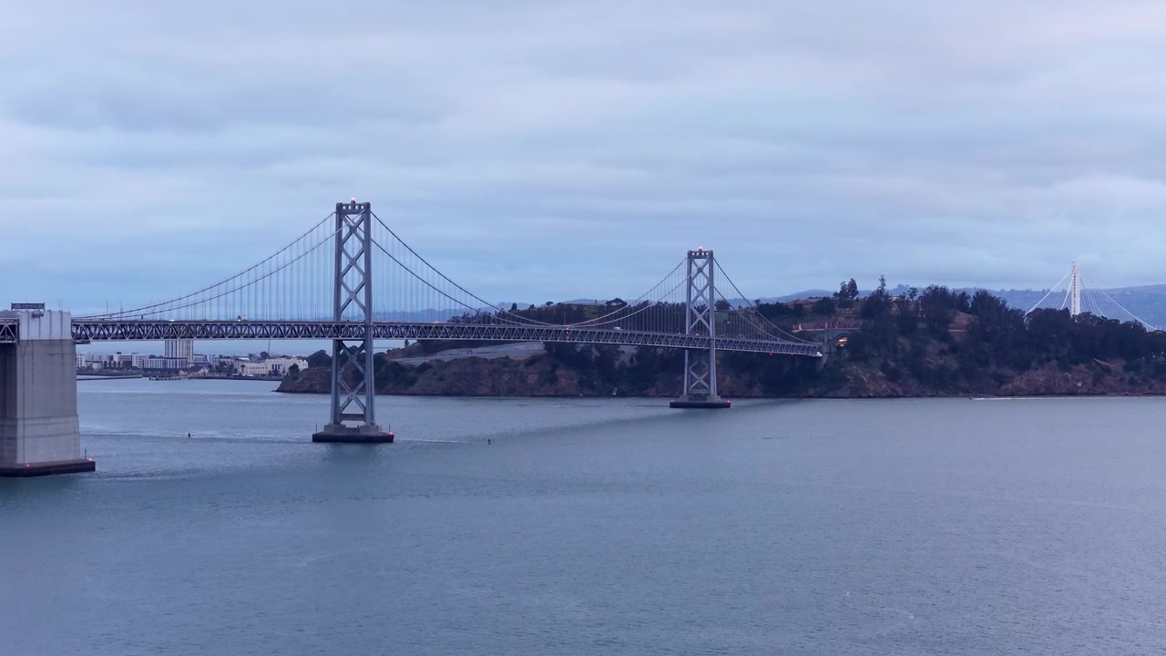 Riprese drone rivelano l'iconico Bay Bridge che attraversa acque blu calme, con lo skyline di San Francisco in lontananza e nuvole serali soffici e suggestive sopra. Inquadratura drone ascendente.