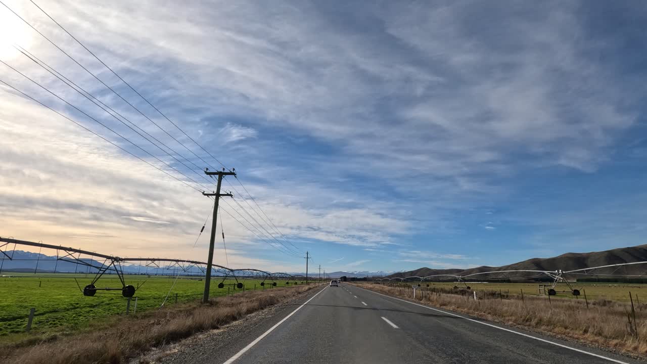 A tranquil drive along a rural New Zealand road, showcasing expansive farmland under a vast, cloudy sky during autumn