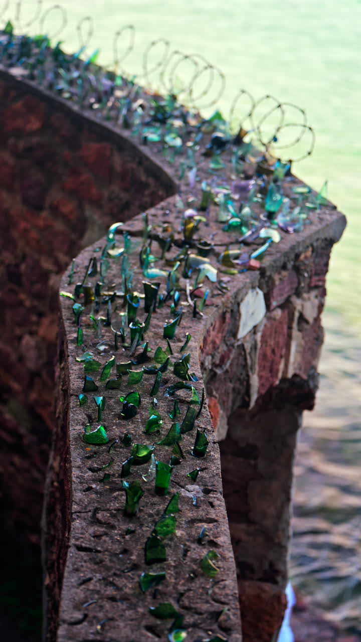 Close up of multiple broken glass shards and barbed wire loops along the edge of a brick wall with a blurred view of the sea. Vertical