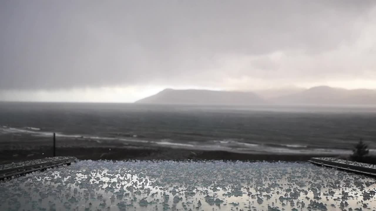 View of stormy sea with dark cloud, falling ice, rough wave, wet shore, and distant hill under cold sky, showing raw power of nature and moody weather along remote coast