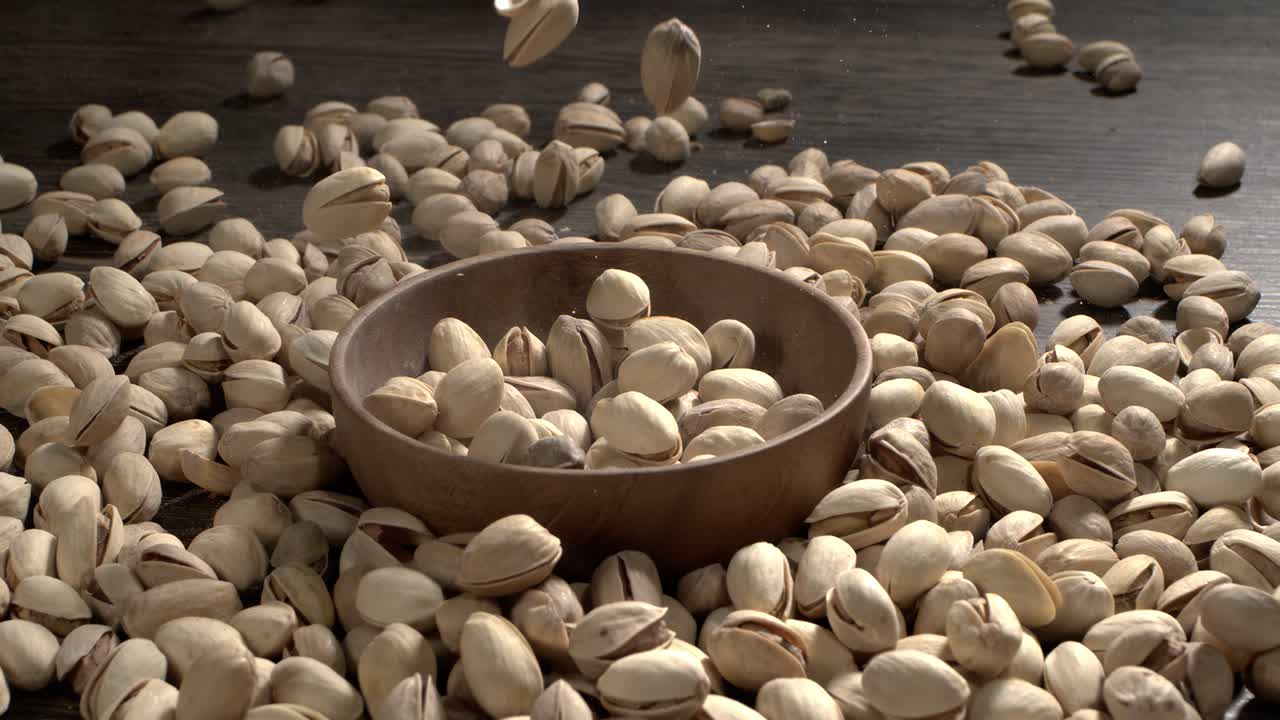 Ciotola di legno piena di pistacchi su tavolo di legno scuro, gustosa frutta secca salata sparsa intorno, concetto di snack salutare e cibo naturale. Scatto con telecamera super slow motion a 1600 fps