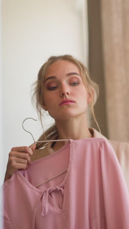 Young confused woman tries on elegant pink and classic black cocktail dresses against mirror choosing fashionable outfit for evening event slow motion