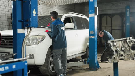 automobile engineers working on white jeep in garage, one fixing wiper, other inspecting back tire with torch light, surrounded by equipment in workshop, performing vehicle maintenance