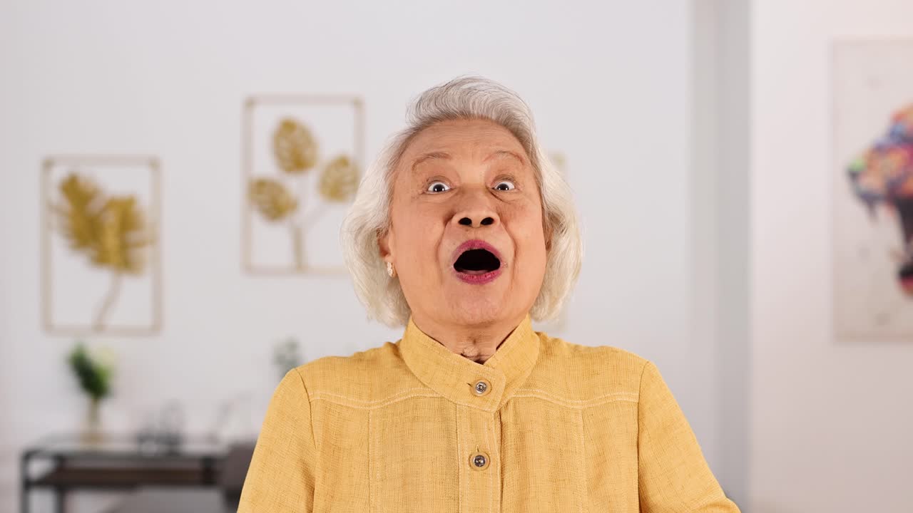 Smiling elderly Asian woman in yellow shirt playfully covers face with hands, expressing shyness in a bright, modern Bangkok living room. Soft natural lighting, static camera