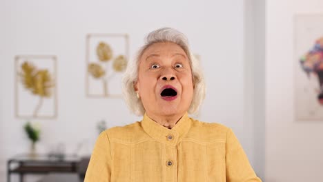 smiling elderly asian woman in yellow shirt playfully covers face with hands, expressing shyness in a bright, modern bangkok living room. soft natural lighting, static camera