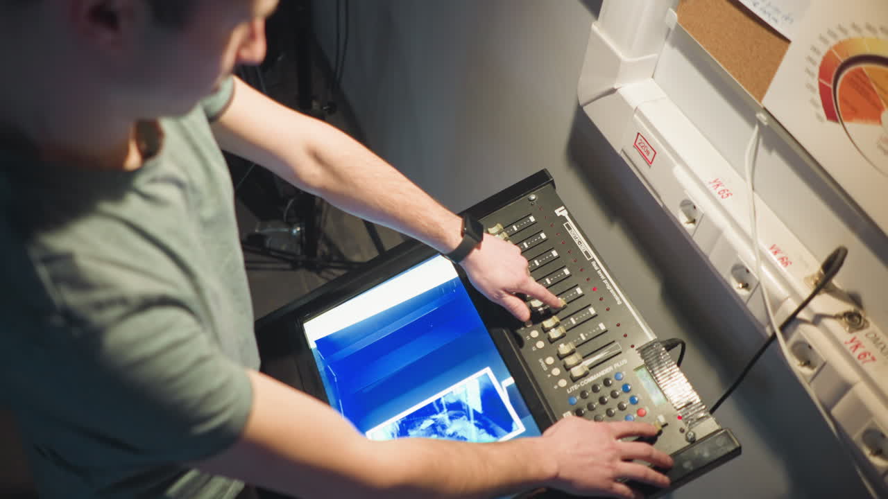 Person interacting with equipment in dark studio setting, adjusting controls on digital panel while viewing display, lighting focused on controls and subject, preparing for task or shoot