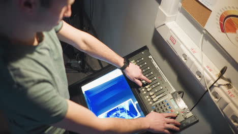 person interacting with equipment in dark studio setting, adjusting controls on digital panel while viewing display, lighting focused on controls and subject, preparing for task or shoot