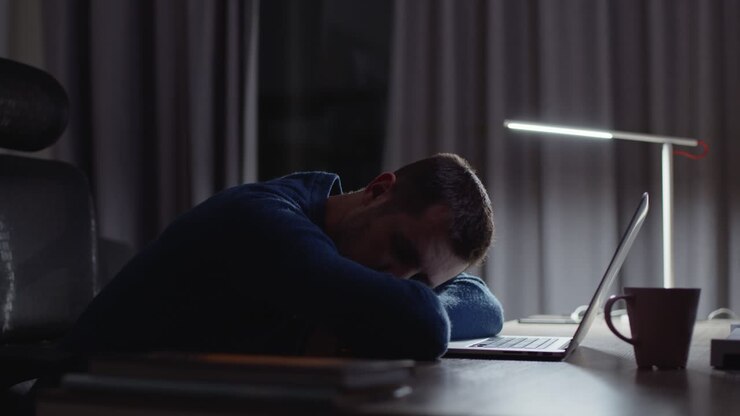 Tired male entrepreneur sleeping in front of laptop due to late night overwork in dark home office workplace
