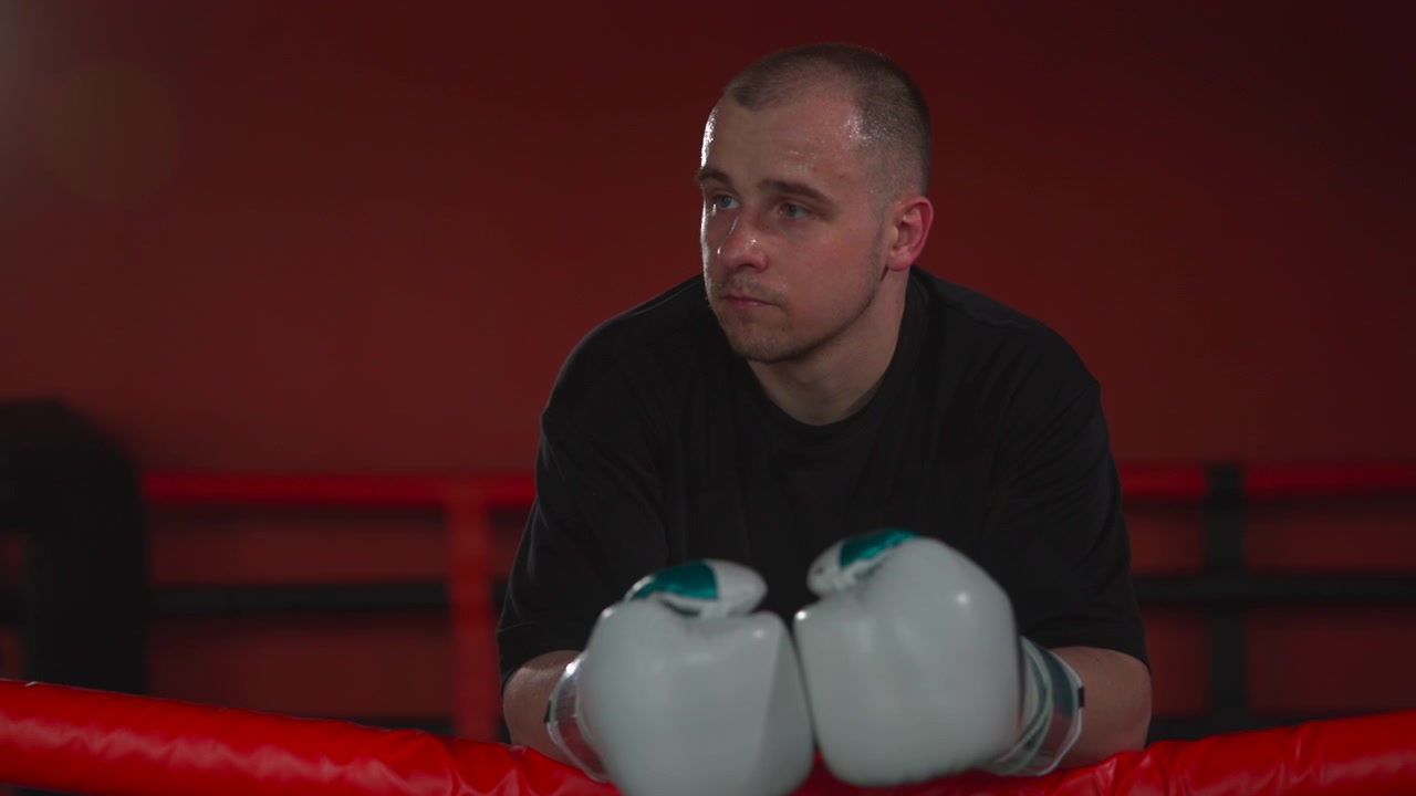 Young male boxer leans over red ring ropes with white gloves together, head lowered, sweat visible on face, dark shirt, short hair, deep red gym background, cinematic low-key lighting, strong