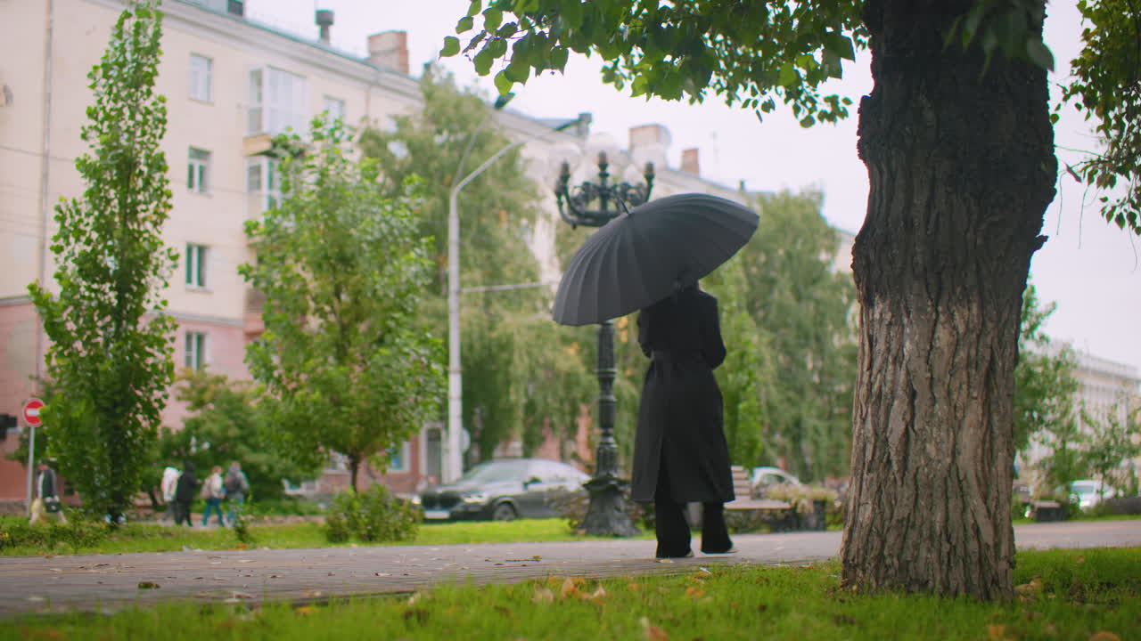 Woman in long black trench coat walking under open umbrella on park pathway, surrounded by green trees and city buildings, overcast weather atmosphere, autumn leaves scattered on ground, urban lifestyle