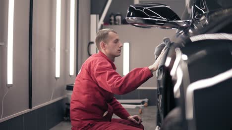 man in red worksuit rubs black expesive car with microfiber wiper. polishing at autocenter.