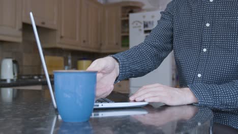 smartly dressed man taking a sip of a hot drink and using track pad on laptop on a kitchen counter while working from home