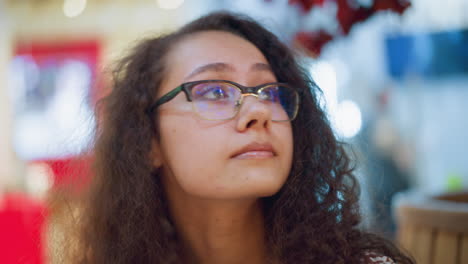 mujer con cabello rizado y gafas en un centro comercial bien iluminado, mirando a su alrededor con una expresión reflexiva, el fondo muestra luces bokeh suaves, creando un ambiente interior vibrante y cálido