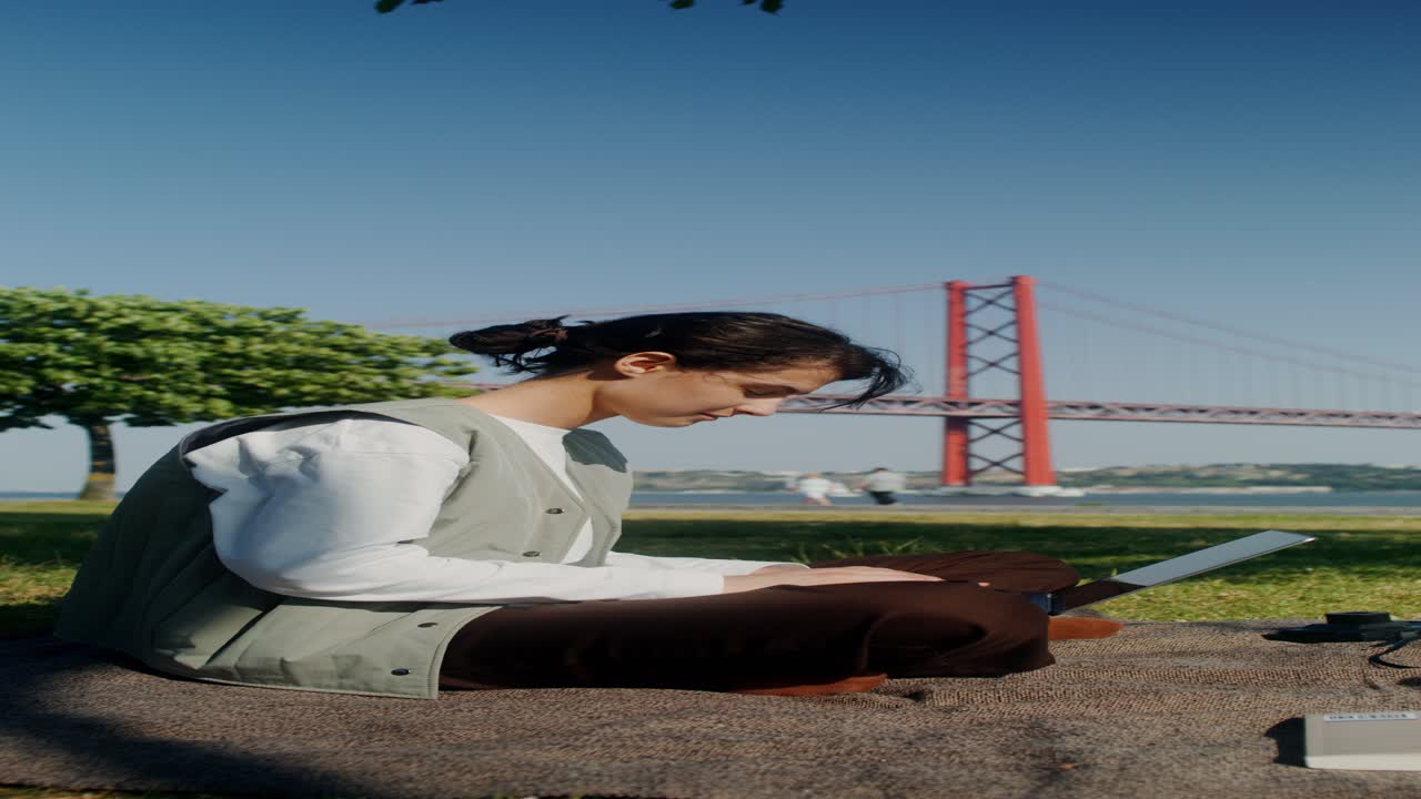 Woman Working on Laptop in a Park with a Bridge in Background