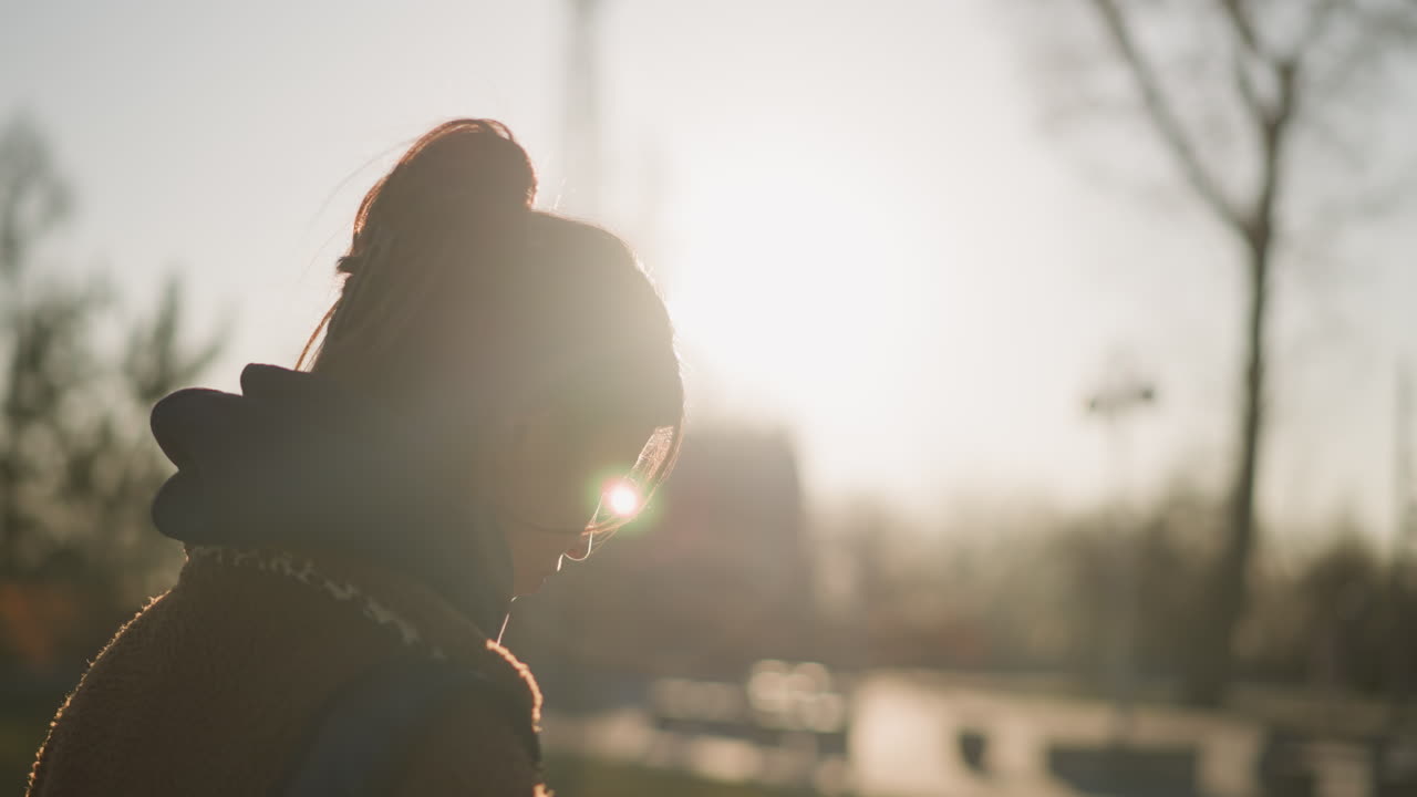 una chica caminando con una expresión de tristeza, su cabello cubriendo parcialmente su cara. el suave resplandor de la puesta de sol en el fondo agrega un estado de ánimo sombrío y reflexivo