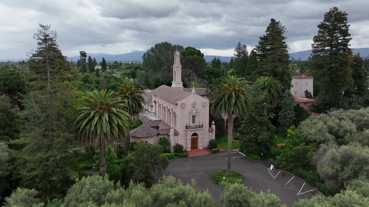 Historisk kyrka med klocktorn reser sig bakom täta olivträd och palmkantad allé i Santa Clara, med berg och dramatiska stormmoln i bakgrunden. Kalifornien, USA. Flygfoto med drönare som närmar sig.