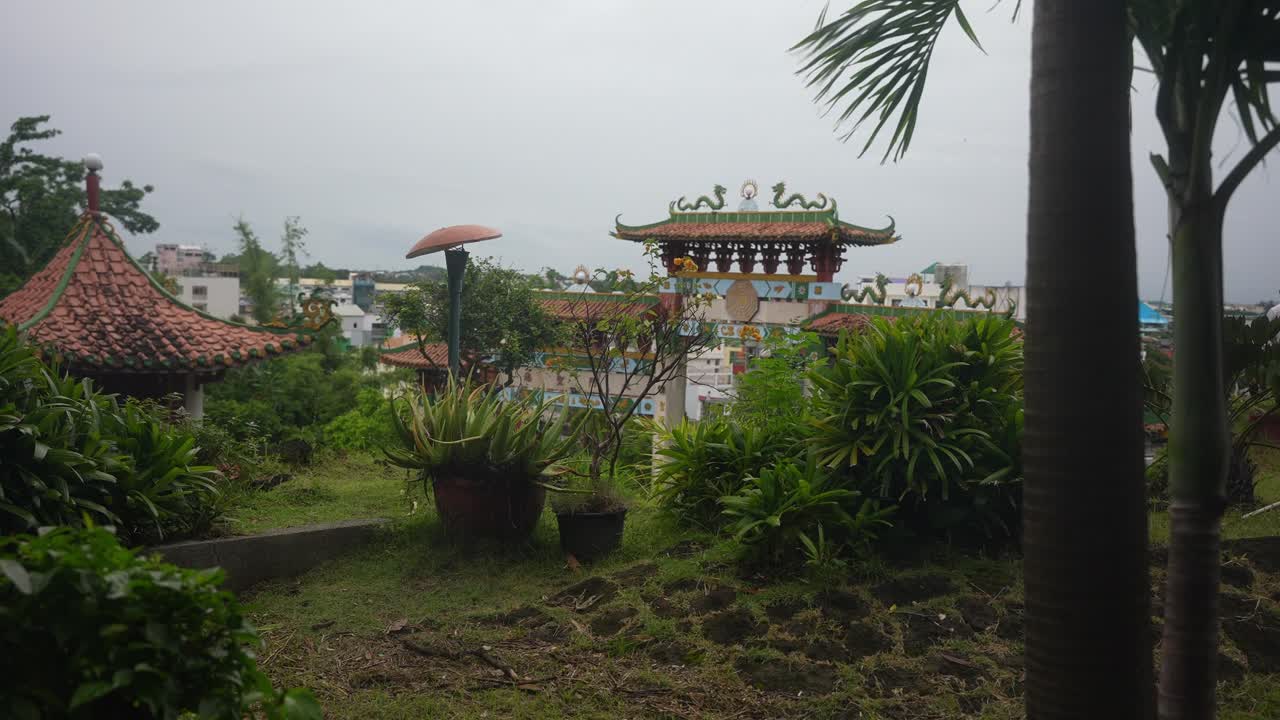A medium static angle holds throughout, framing a temple facade with green and red tiled roofs, dragon sculptures, and geometric motifs. The lush garden and overcast sky evoke calm