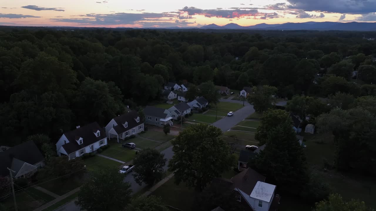 Quiet American suburban neighborhood at sunset. Rows of houses line the street, surrounded by trees, with the Blue Ridge Mountains silhouetted against colorful evening sky. Aerial approaching flight