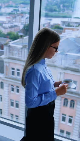 lunch break in the modern office with beautiful view from window. blonde female employee walking up to window with a cup of coffee. high angle view. vertical video
