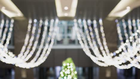 luminous light bulbs are hanging from the ceiling indoors. these decorations are used at christmas. the blurred background is people entering or leaving the building. out of focus. in the center of the screen is a green christmas tree.