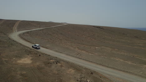 Seitliche-Luftaufnahme,-Die-Einem-Weißen-Auto-Auf-Einer-Vulkanstraße-Auf-Der-Insel-Fuerteventura-An-Einem-Sonnigen-Tag-Folgt