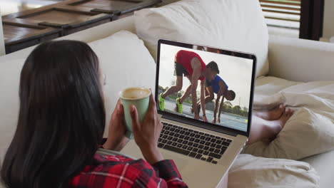 compuesto de una mujer sentada en casa sosteniendo un café viendo un evento de atletismo en una computadora portátil