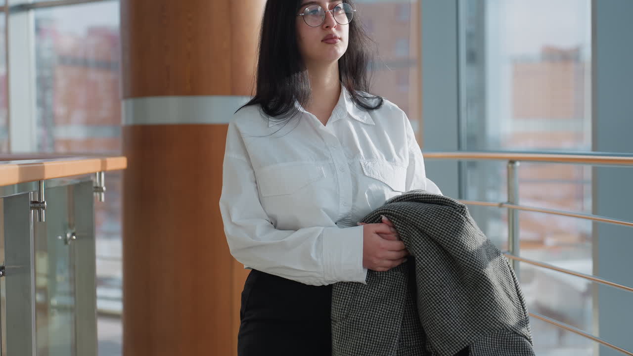 Close up of confident professional in black trousers and white shirt walking through bright mall gallery holding checkered jacket, surrounded by glass panels and sunlit modern architectural elements