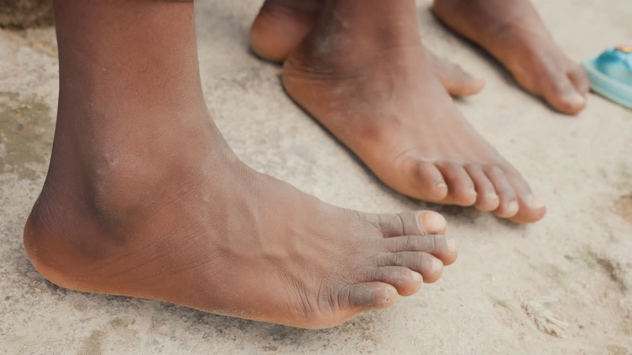 Piede nudo di colore che calpesta terreno sabbioso, primo piano della pianta del piede che si solleva e degli spruzzi di sabbia, energia dinamica e slancio atletico che suggeriscono uno sprint o un calcio, inquadratura ravvicinata con compagni di squadra e illuminazione solare sullo sfondo.