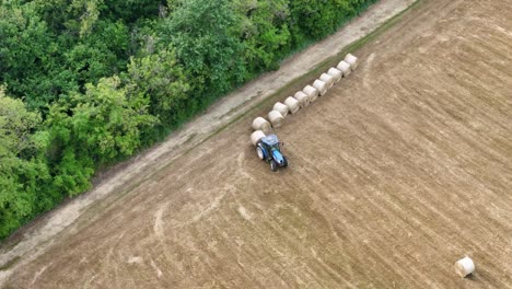Drone shot of a farmer using a blue tractor to align cylindrical hay bales along a wooded border in a dry springtime field near Piacenza, northern Italy, preparing for bale storage transport.