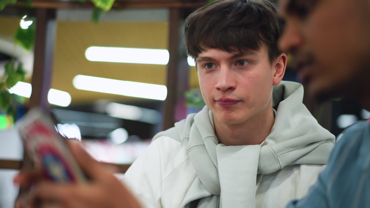 European young man watching friend phone with focused expression in blurred background sharing reaction and conversation casual indoor setting showing curiosity and connection over digital content