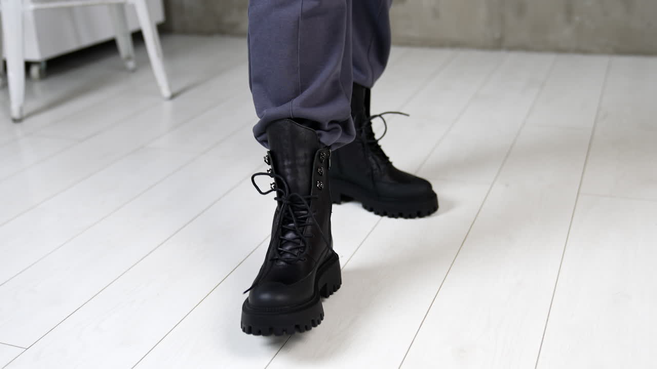 Black massive army boots with laces on model's legs. Woman walks in stylish footwear on the white floor in studio. Top view close up.