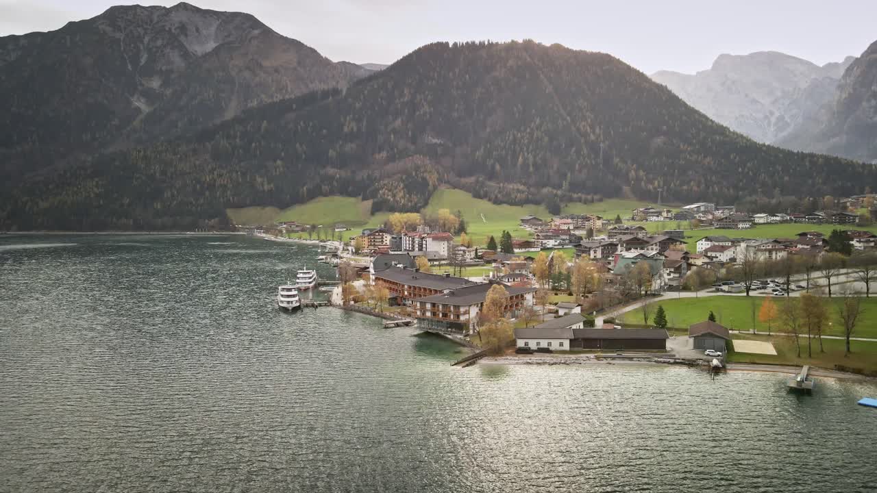 A captivating drone shot from Achensee Pertisau, flying outward to reveal the picturesque lake town surrounded by mountains and vibrant turquoise waters. A serene alpine scene in Austria.