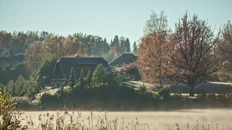 shot of morning fog passing by in timelapse over rural cottages and river alongside lush green vegetation on a sunny morning