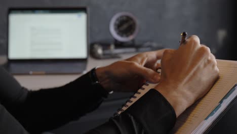 Close-up-view-of-a-man-writing-lyrics-in-a-notebook-with-a-laptop-in-the-background