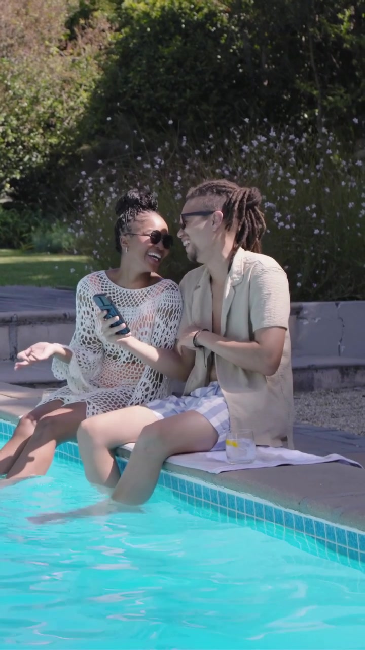 Video vertical: Levantando el teléfono, pareja apoyada en la piscina, mujer con pareo mirando y brindando con copas.