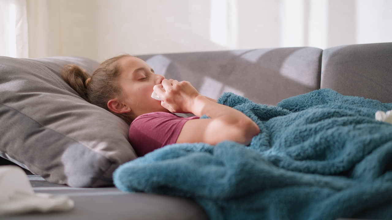 Visuale laterale di una giovane ragazza sdraiata, che starnutisce in un fazzoletto con una coperta addosso, indisposta e stanca, con fazzoletti di carta sparsi intorno, a indicare i sintomi dell'influenza, mentre si riposa in un soggiorno accogliente
