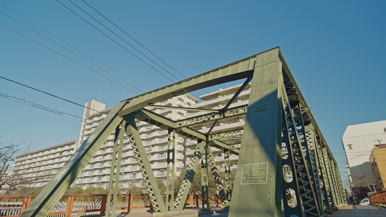 Fukuju Bridge's yellow-green steel truss fills the frame against vivid blue sky in Tokyo, with a large residential building behind, captured with a tilt-down camera movement revealing the full structu