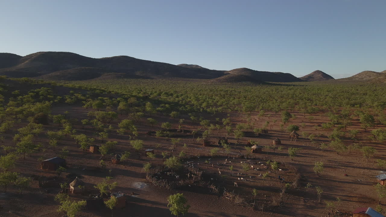 Drone flies over a Himba tribe camp in Namibia during golden hour, revealing red earth, traditional huts, scattered trees, and distant mountains in stunning light.