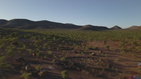 Drone flies over a Himba tribe camp in Namibia during golden hour, revealing red earth, traditional huts, scattered trees, and distant mountains in stunning light.