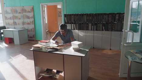 college student in casual clothing and cap sitting at desk in bright library, writing notes from open book with several stacked books nearby, sunlight streaming across wooden floor