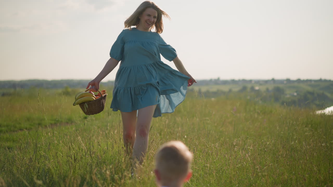 A woman in a flowing blue dress joyfully runs through a vibrant,grassy field toward two young boys in white tops.One boy raises his hand to welcome her as she laughs and carries a basket full of fruit