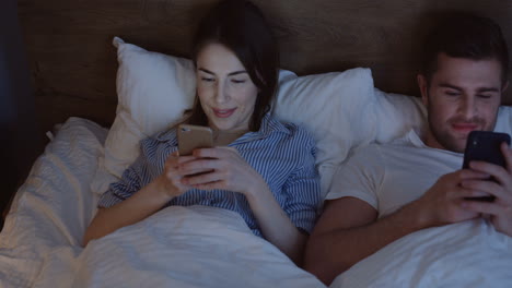 vista superior de una joven caucásica y un hombre acostados en la cama, charlando y riendo en el teléfono inteligente a altas horas de la noche