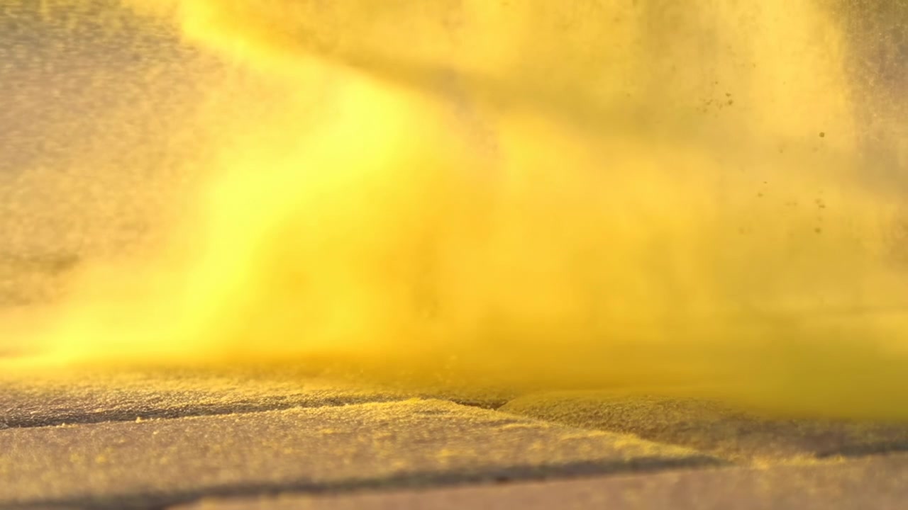 Densa nuvola di polvere gialla si deposita e si diffonde su un pavimento ruvido dopo un disturbo, formando una foschia di particolato semi-opaca con fini particelle sospese sopra il terreno strutturato e diffusa da luce calda.