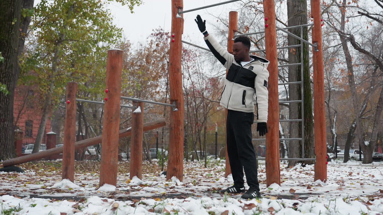 젊은이가 야외 겨울 운동장에서 유연성을 위해 팔을 펴고, 따뜻한 털과 장갑을 입고, 나무와 눈 인 땅과 함께 나무 피트니스 장비 근처에서 운동합니다.