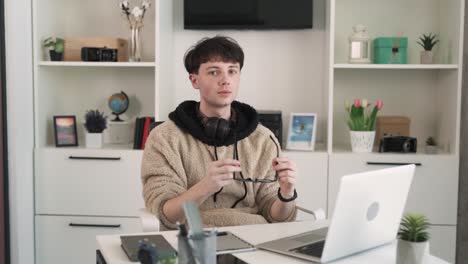 there is a portrait of a young man sitting at a desk in an office, in front of a laptop. he puts on spectacles and looks attentively into the camera. the man is seated in a modern, bright office.