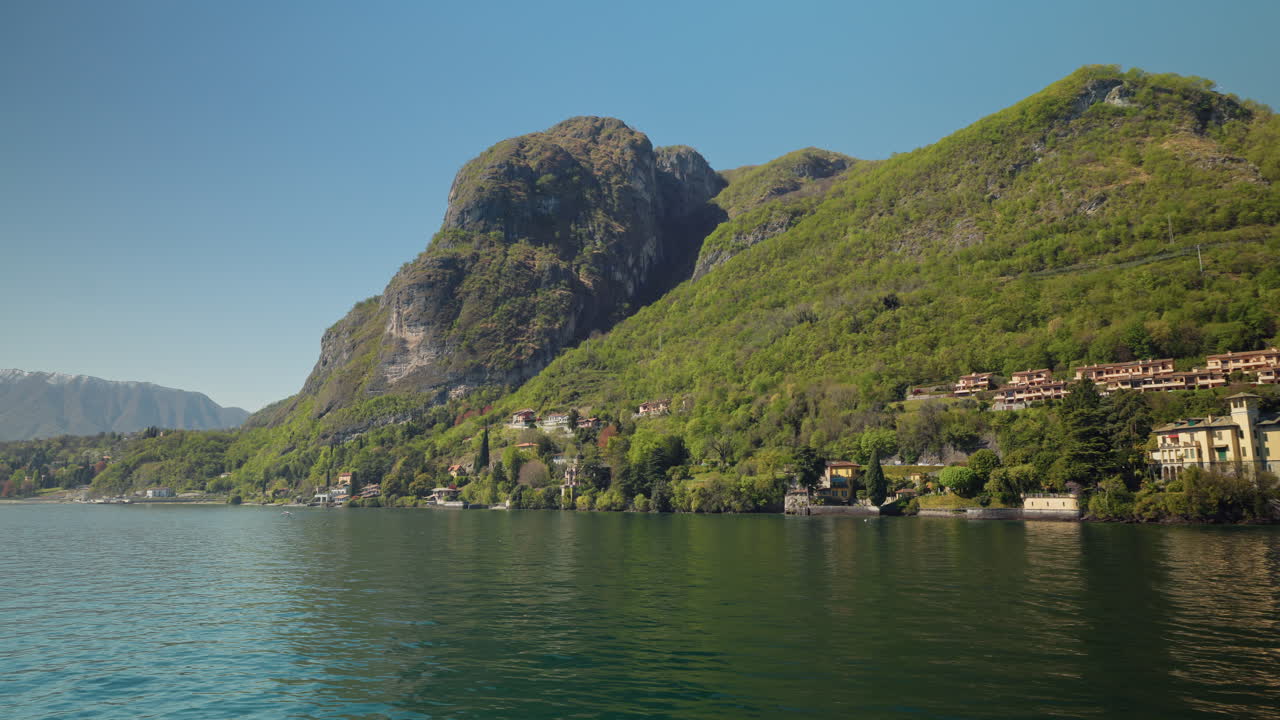 Una ripresa sull'acqua in movimento cattura una drammatica scogliera rocciosa calcarea che si erge sopra rigogliose colline primaverili punteggiate da ville e giardini italiani che digradano verso le rive turchesi del Lago di Como in Italia.