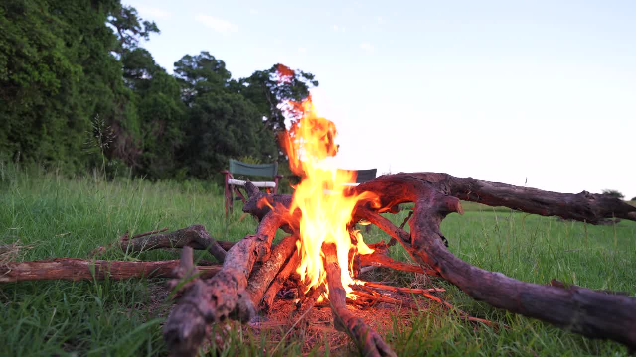 Warm bonfire flames flicker gently just before sunset at a remote campsite in the wild bush of Maasai Mara, Kenya. A serene, atmospheric shot capturing adventure, warmth, and wilderness