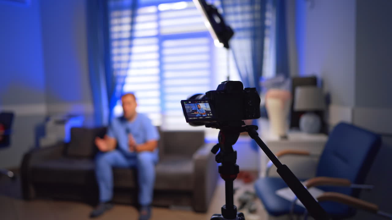 Camera set on tripod recording a video. Man wearing blue uniform sits at the sofa talking. Selective focus